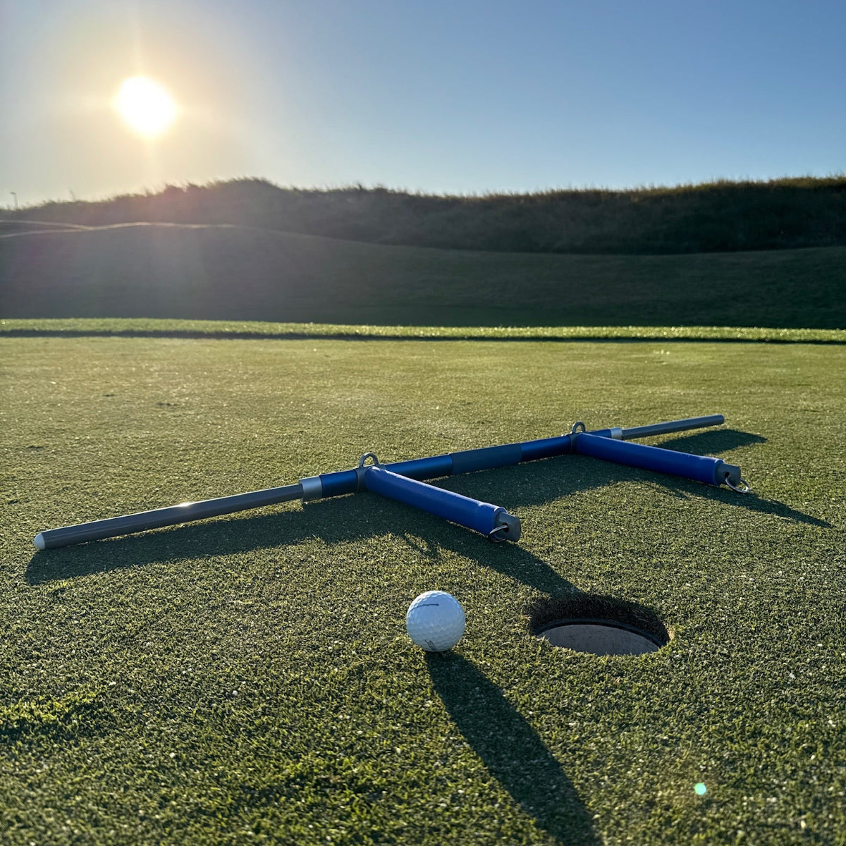 Golf ball next to a the putt rocker on a golf course with a hole in the foreground and sun in the background.